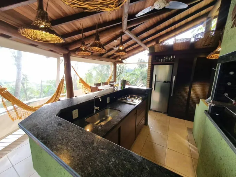 Espaço gourmet com bancada de granito, cooktop, geladeira, churrasqueira e redes com vista para o bosque