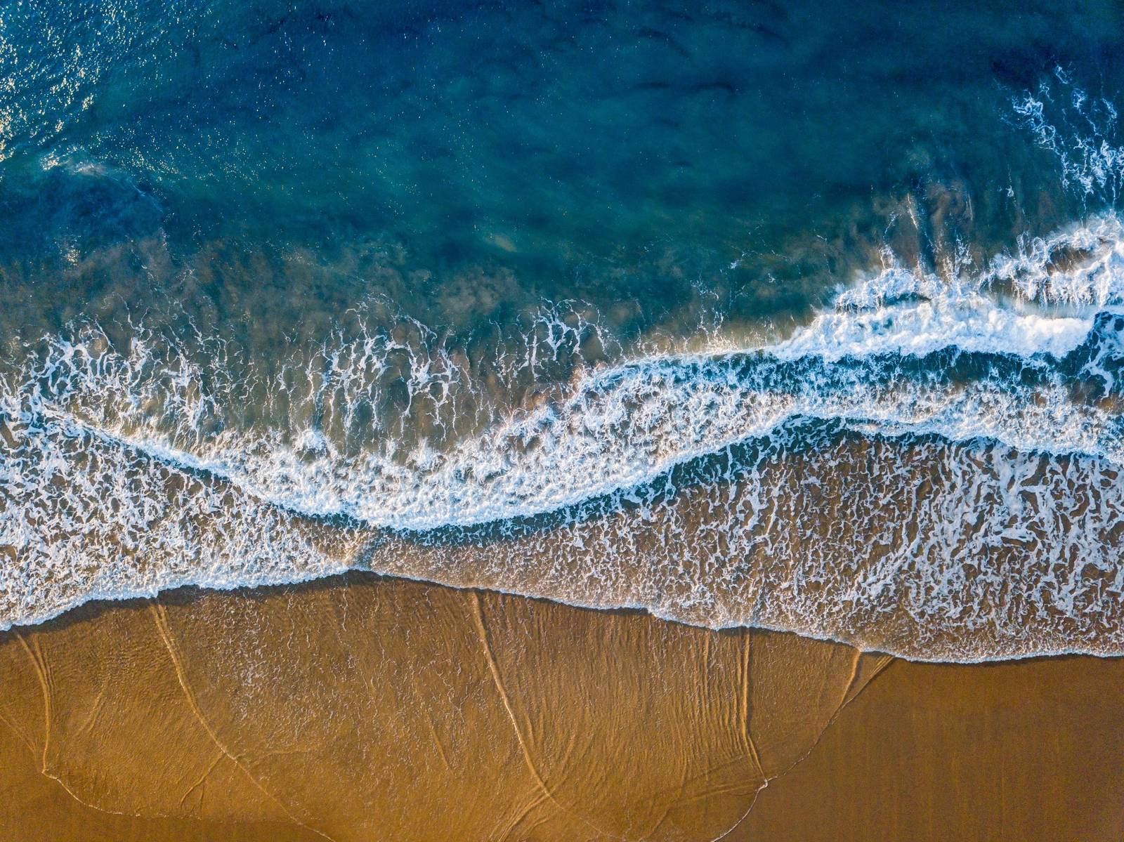Vista aérea das ondas chegando à areia dourada da praia, a 600 m do Refúgio do Ermitão