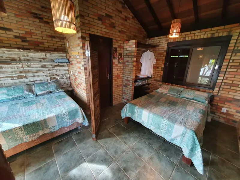 Suíte 02 com duas camas de casal, paredes de tijolo à vista e luminárias de palha